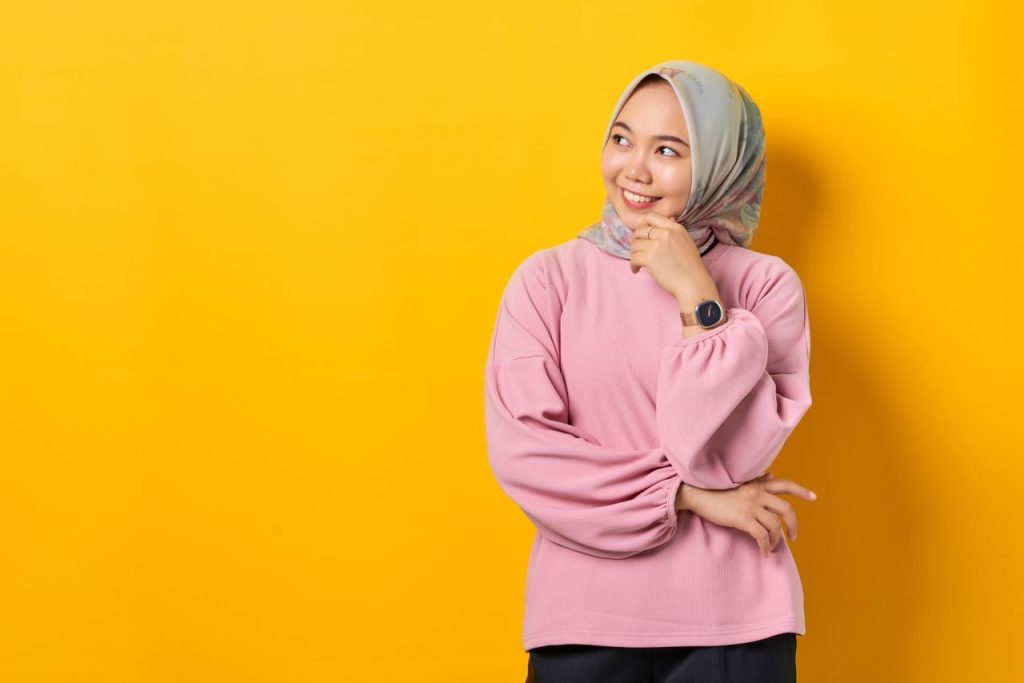 Asian woman in hijab smiling thoughtfully against a vibrant orange background.
