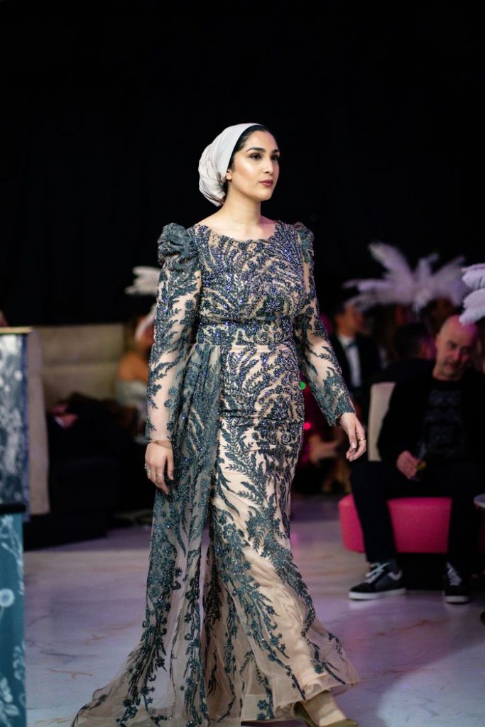 Model walking on the runway in an ornate embroidered dress and headscarf.